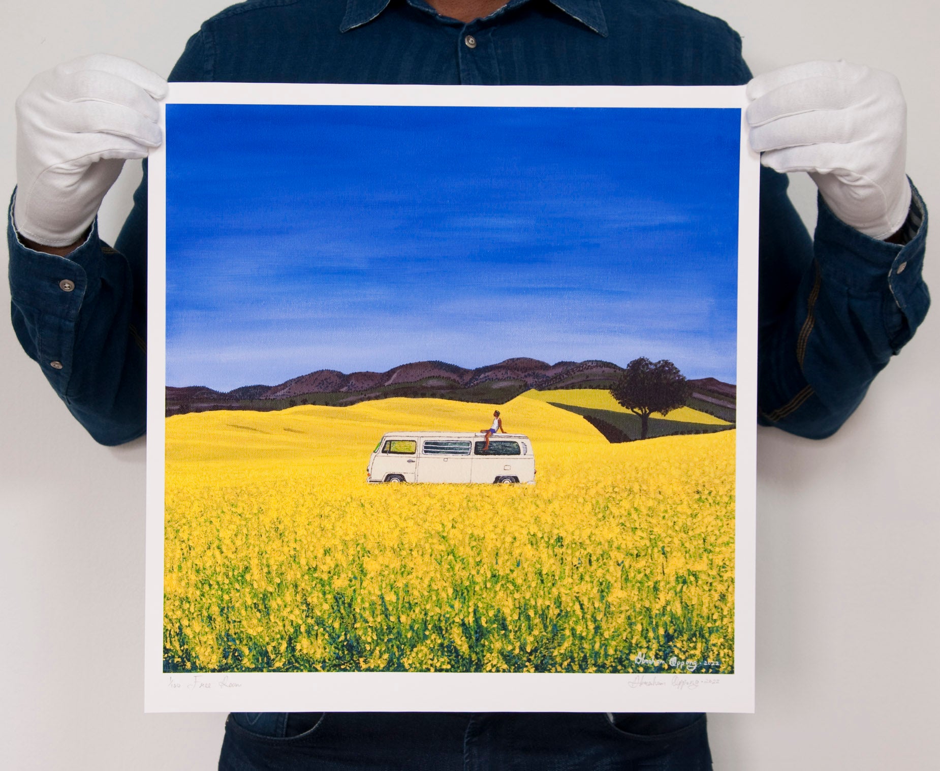 Artist holding high quality giclee print of free roam, a man sitting on the roof of a white campervan on a sunny day of blue skies in yellow flower fields with trees and hills in the background. Modern high quality affordable art prints. 